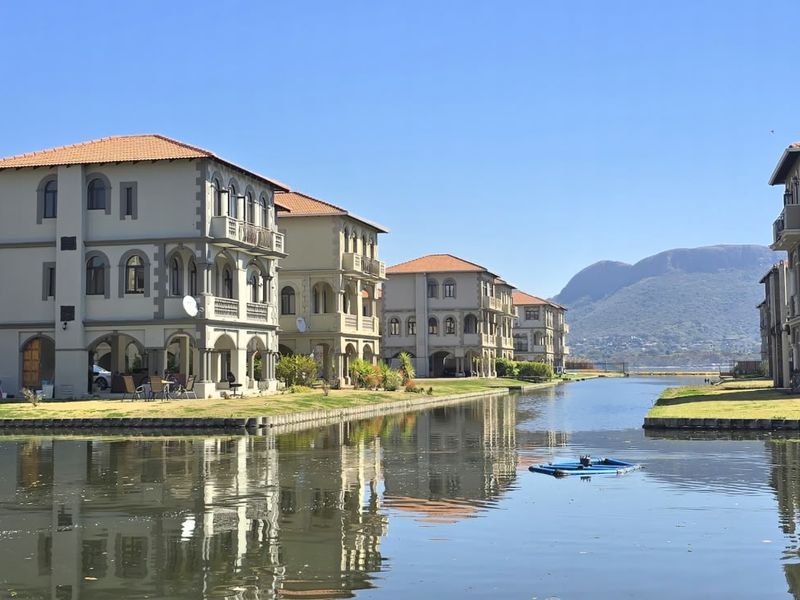 A scenic view of multi-story residential villas situated along a canal, featuring architectural arched balconies and well-maintained lawns with mountain views.