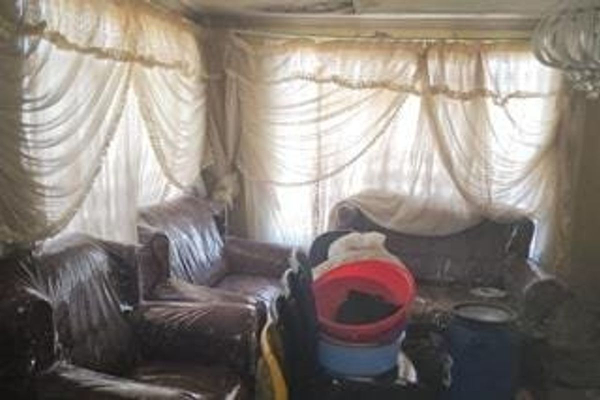 A living room space featuring large windows with heavy curtains and visible ceiling damage, requiring significant renovation and modernization.