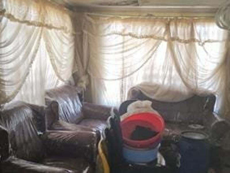 A living room space featuring large windows with heavy curtains and visible ceiling damage, requiring significant renovation and modernization.