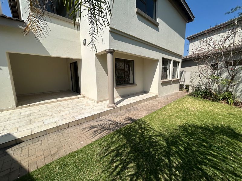 A rear view of the property featuring a covered tiled patio, paved walkway, and a well-maintained lawn area.