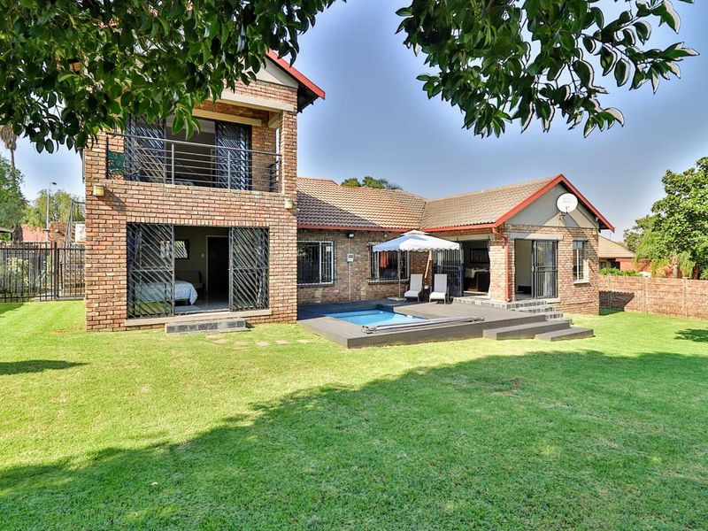 Rear view of a double-story face brick house featuring a swimming pool, timber deck, and an upper-level balcony overlooking a spacious garden.
