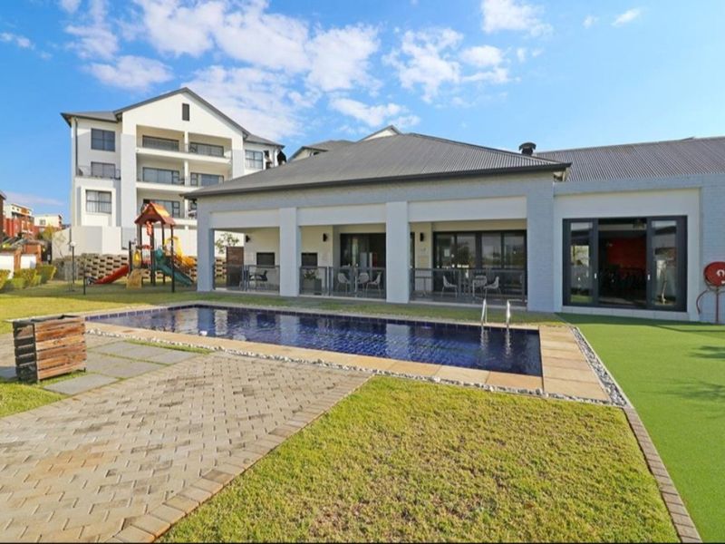 A well-maintained communal pool area featuring a swimming pool, paved surrounds, artificial lawn, and a covered patio area attached to the clubhouse building.