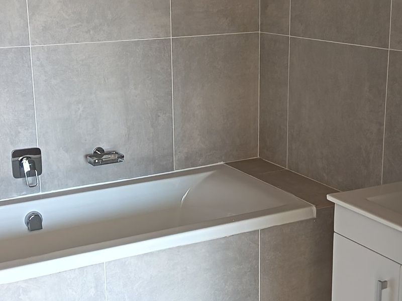 A modern, clean bathroom featuring a built-in bathtub with wall-mounted fixtures, neutral grey tiling, and a white vanity cabinet.