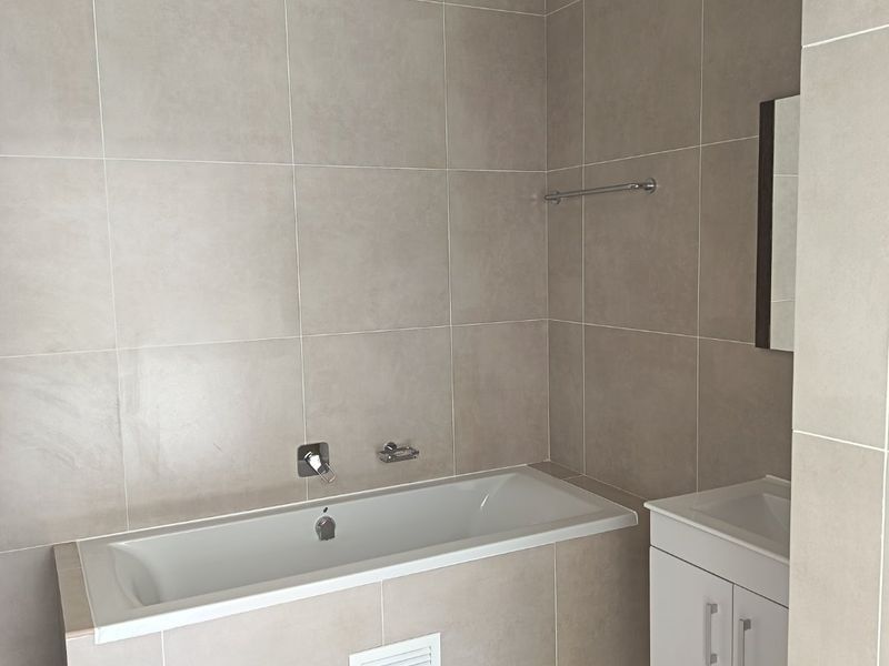A clean, modern bathroom featuring neutral floor-to-ceiling tiling, a built-in bathtub with wall-mounted fixtures, and a white vanity cabinet with an integrated basin.