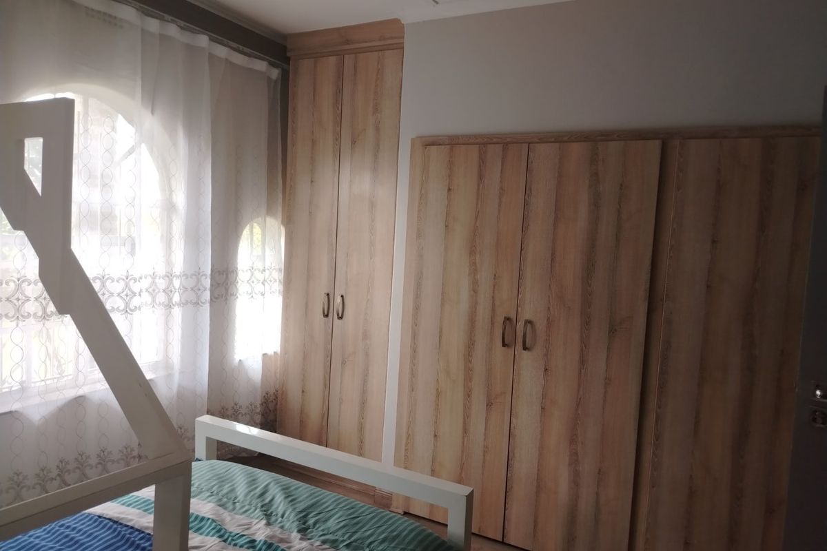 A bedroom featuring light wood-grain built-in cupboards, laminate flooring, and an arched window with sheer curtains.