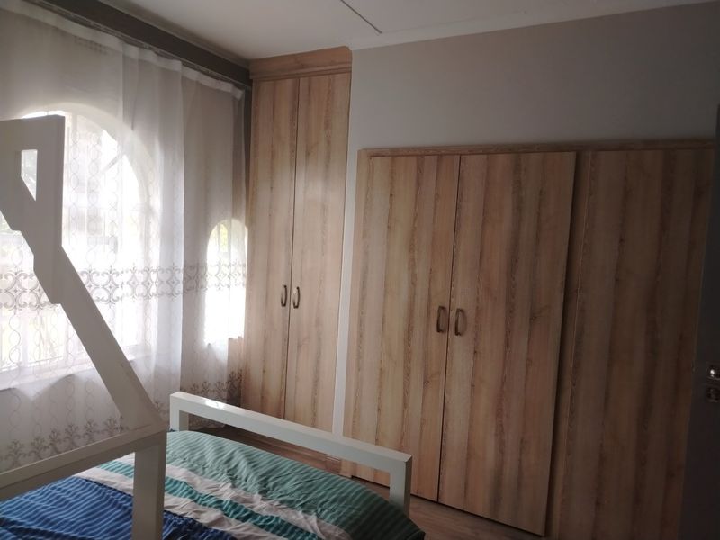A bedroom featuring light wood-grain built-in cupboards, laminate flooring, and an arched window with sheer curtains.