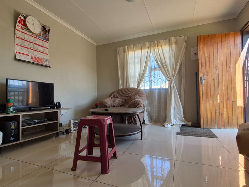 A living room featuring tiled flooring, a wooden entrance door, and a window fitted with security bars.