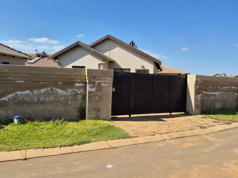 Front view of the property featuring a secure sliding gate, paved driveway, and a perimeter wall with a tiled roof house visible in the background.