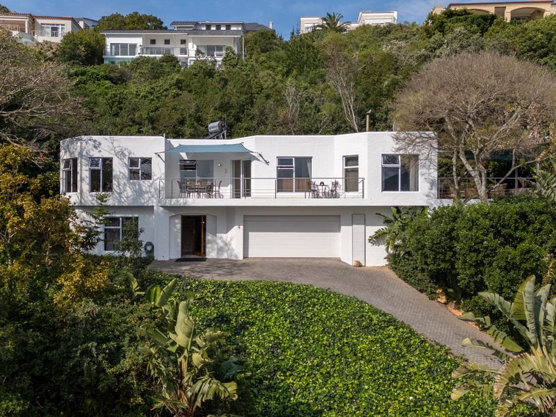 A multi-level white residential property featuring a paved driveway, integrated double garage, and an upper-level balcony set against a lush, elevated hillside.