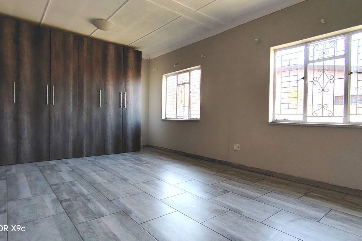 A spacious, empty bedroom featuring modern wood-look floor tiles, built-in cupboards, and windows fitted with security bars.
