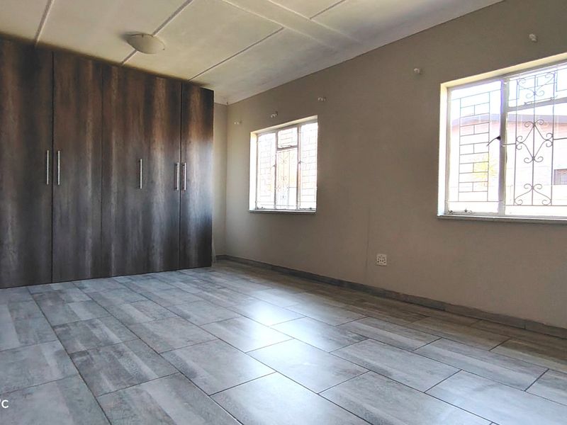 A spacious, empty bedroom featuring modern wood-look floor tiles, built-in cupboards, and windows fitted with security bars.