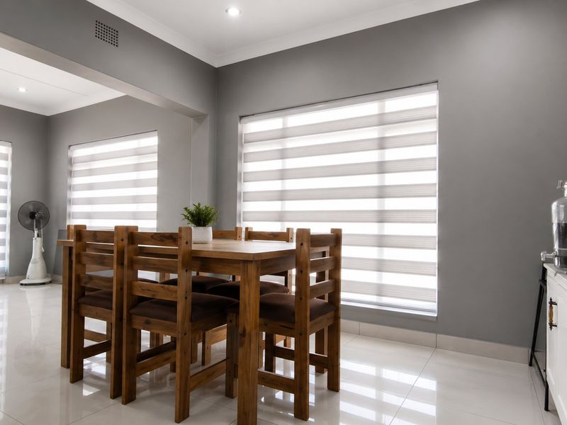 A bright and modern dining area featuring neutral grey walls, polished porcelain floor tiles, and contemporary zebra blinds.