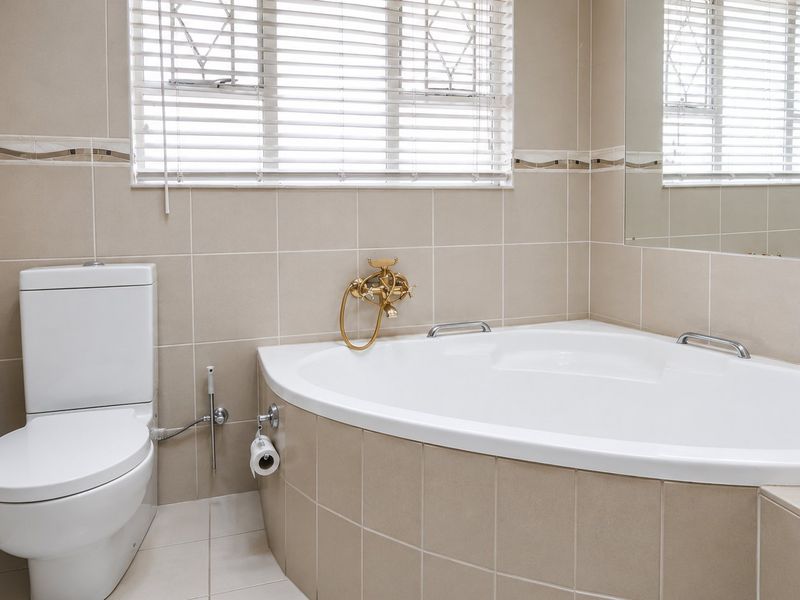 A clean, neutral-toned bathroom featuring a corner bathtub, a modern toilet, and large wall-mounted mirror with tiled finishes throughout.