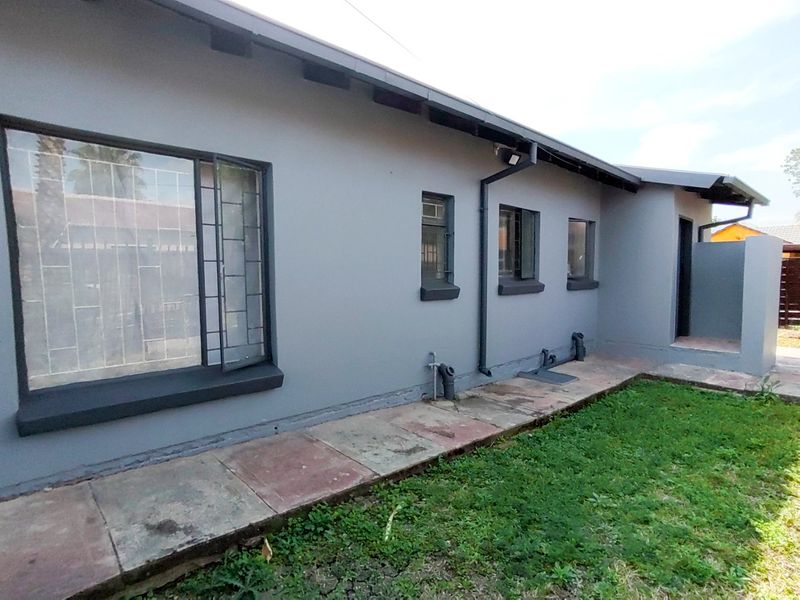 Rear exterior view of the house featuring grey painted walls, security bars on windows, and a paved walkway alongside a small lawn area.