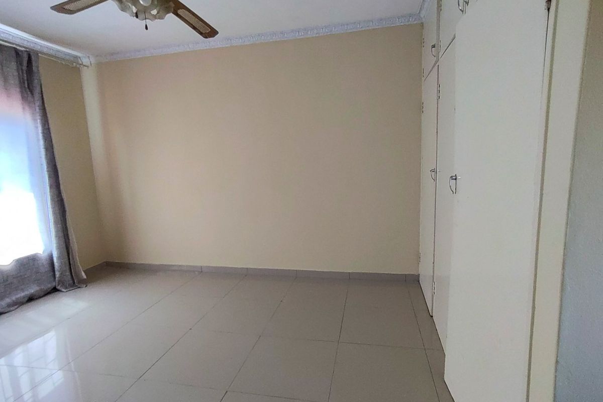 A bright, empty bedroom featuring neutral tiled flooring, built-in cupboards for storage, and a ceiling fan.