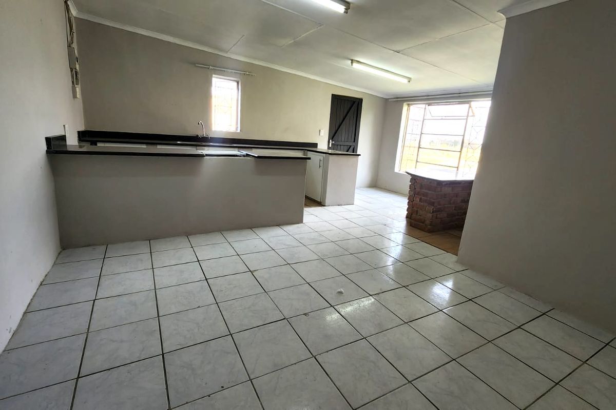 An empty, open-plan kitchen area featuring tiled flooring, built-in cabinetry, a sink, and a unique brick breakfast bar, ready for renovation.