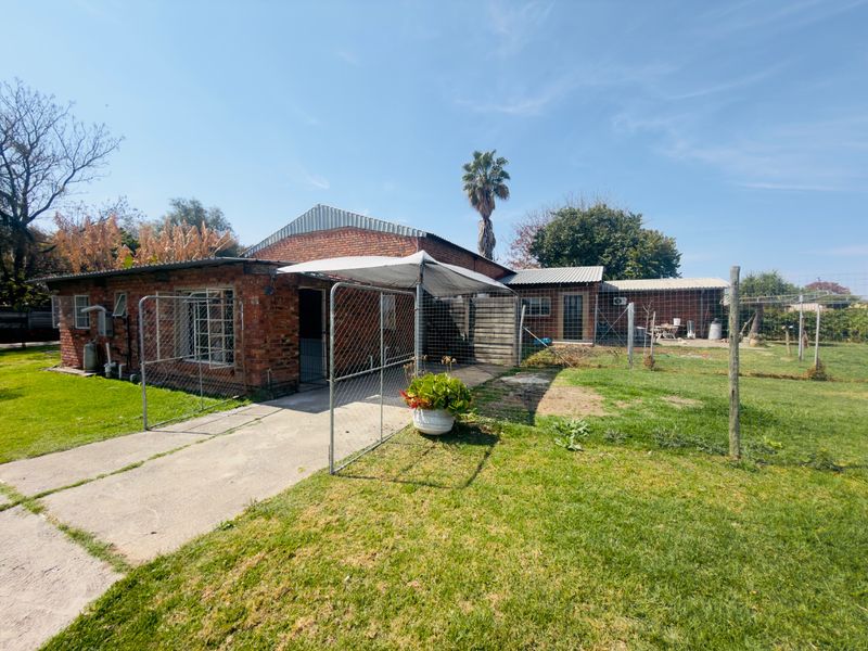 Rear view of a single-story brick house featuring a corrugated metal roof, concrete pathways, and a large garden area with perimeter fencing.