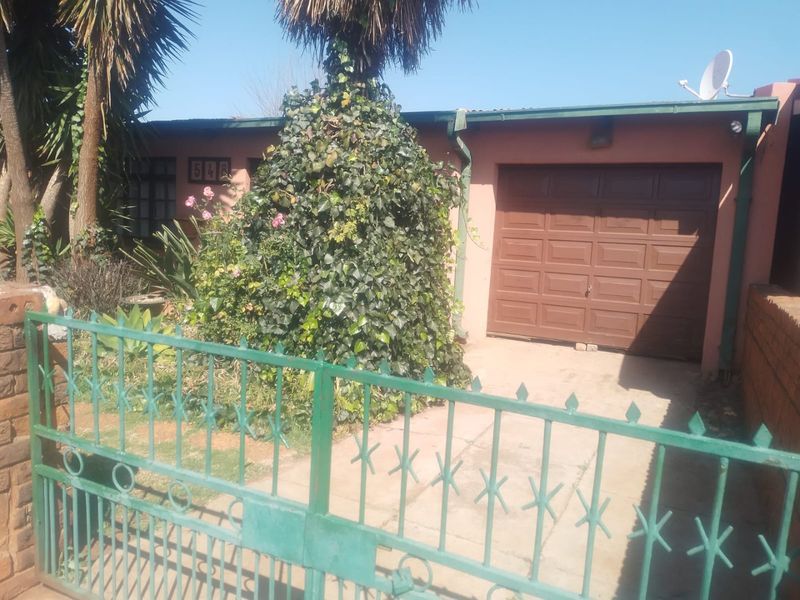Front elevation of the property featuring a metal gate, concrete driveway, and a single garage door, partially obscured by mature garden vegetation.