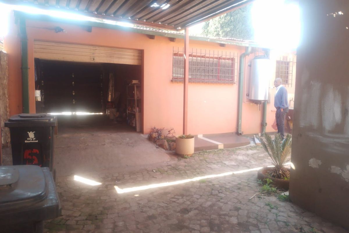 Rear exterior view showing a garage, paved courtyard area, and external geyser installation with security bars on windows.