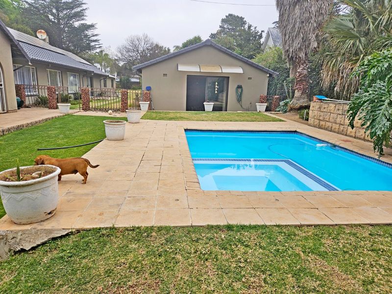 A well-maintained outdoor pool area featuring a paved deck, manicured lawn, and a detached outbuilding with sliding glass doors.