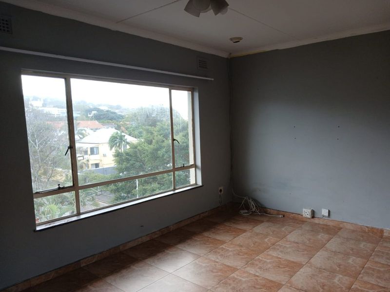An empty living room featuring large windows with neighbourhood views, ceramic tiled flooring, and grey painted walls requiring minor cosmetic attention.