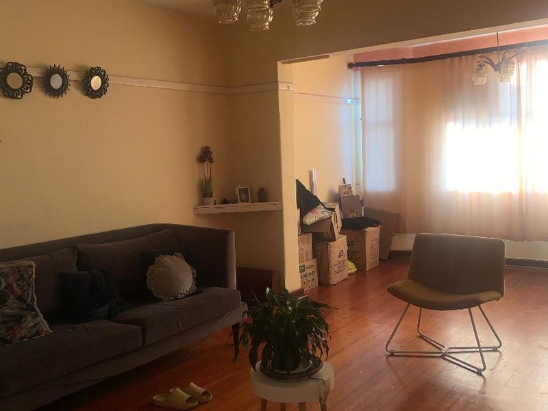 A living room featuring original hardwood flooring and a vintage ceiling light fixture, currently showing signs of ceiling plaster damage and wear.