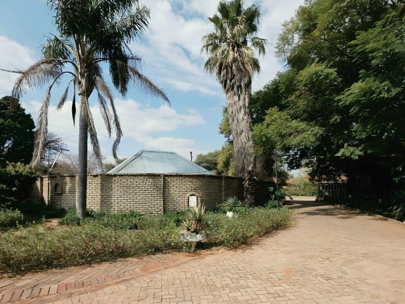 A view of the property entrance featuring a paved driveway, mature palm trees, and a brick boundary wall with a corrugated metal roof structure.