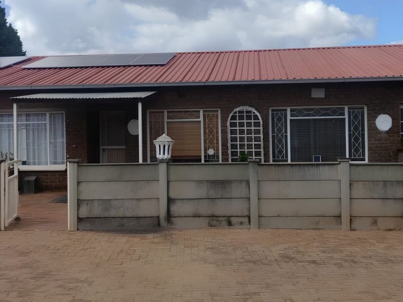 Front elevation of a single-story brick house featuring a corrugated metal roof with solar panels, a paved driveway, and a precast concrete boundary wall.