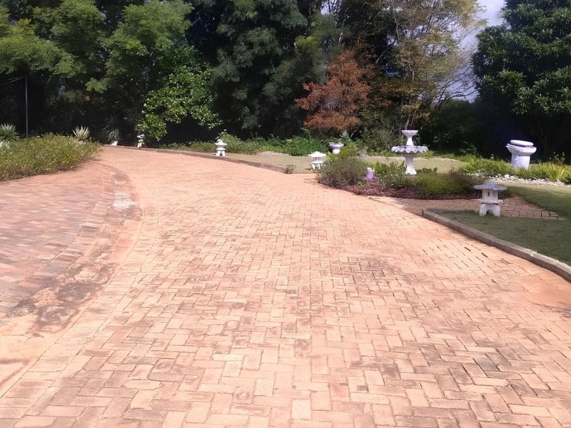 A spacious garden area featuring a long brick-paved driveway surrounded by established trees, lawn, and decorative stone garden ornaments.