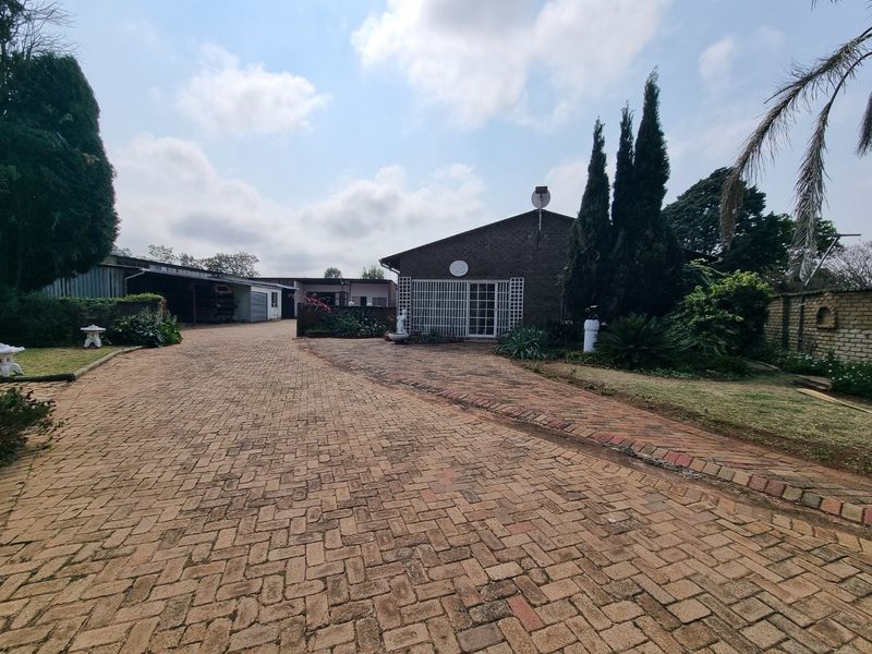 Front view of the property featuring a large brick-paved driveway, established garden, and a covered carport area.
