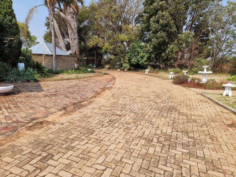 A wide brick-paved driveway leading through a mature garden towards a brick outbuilding on the property.