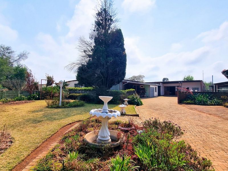 Spacious rear exterior featuring a large brick-paved driveway, established garden with a central fountain, and access to outbuildings.