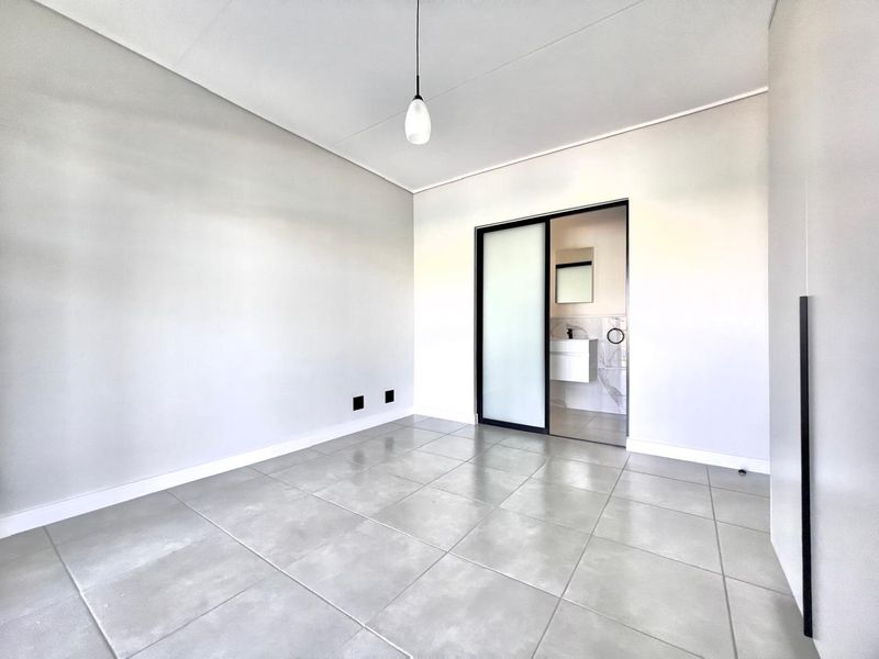 A bright, modern bedroom featuring neutral tiled flooring, a sleek pendant light, and a sliding door providing access to an en-suite bathroom.