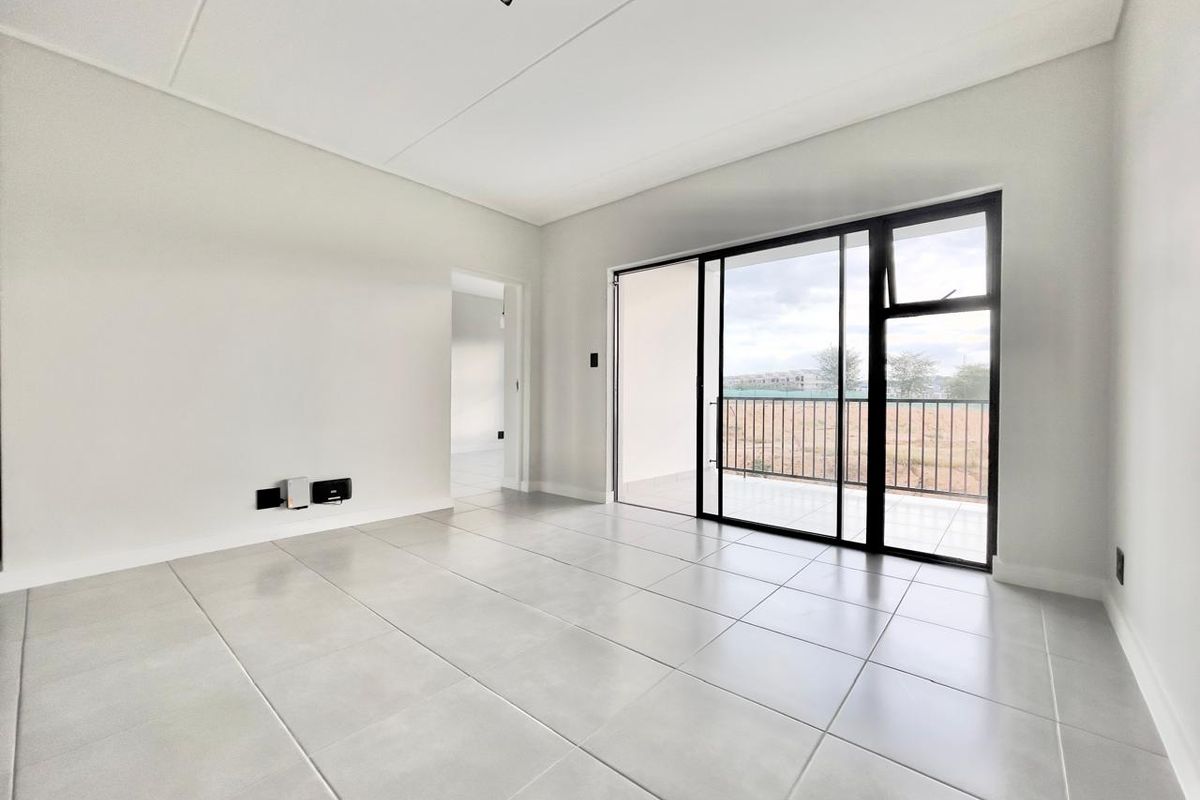 A bright, modern, and empty living room featuring large format floor tiles, track lighting, and a sliding glass door leading to a private balcony.