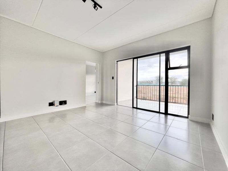 A bright, modern, and empty living room featuring large format floor tiles, track lighting, and a sliding glass door leading to a private balcony.