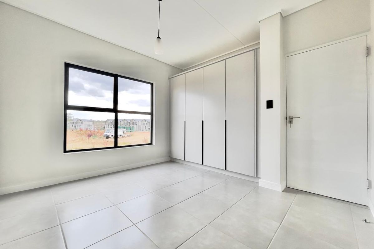 A bright, modern, and empty bedroom featuring neutral tiled flooring, built-in cupboards, and a large window with views of the surrounding development.