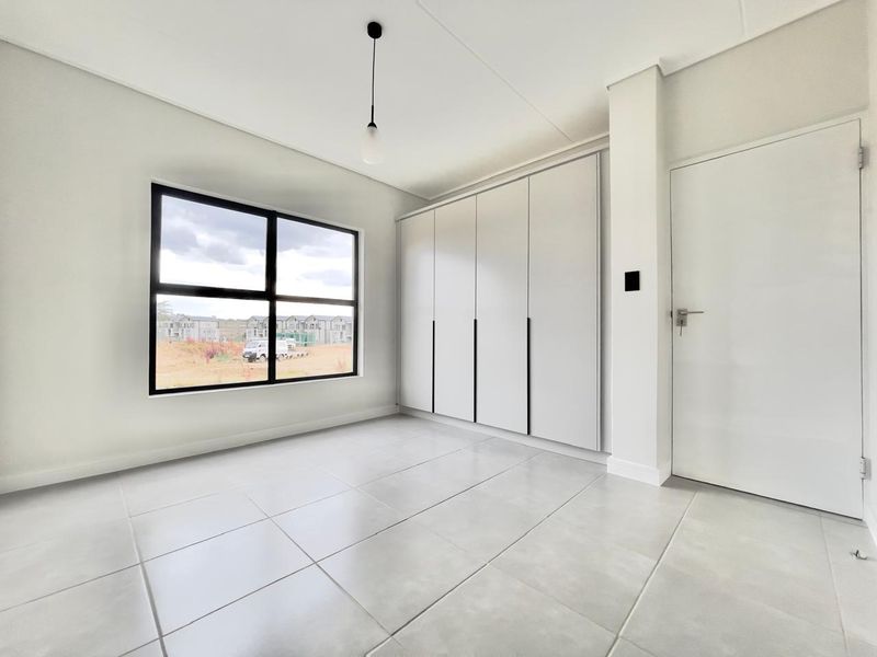 A bright, modern, and empty bedroom featuring neutral tiled flooring, built-in cupboards, and a large window with views of the surrounding development.