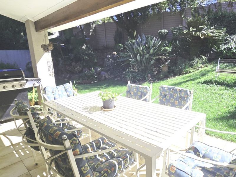 A covered outdoor patio area featuring tiled flooring, overlooking a landscaped garden with a retaining wall.