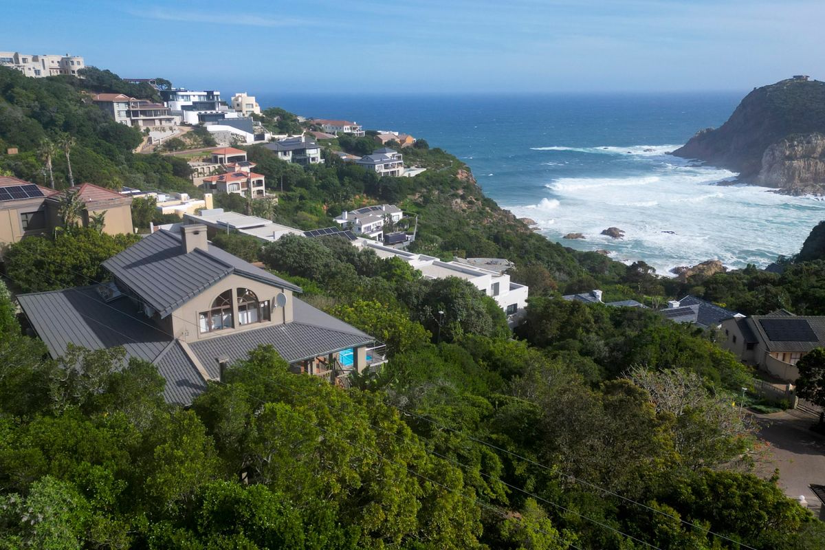 An elevated aerial view of a coastal residential estate featuring homes with modern pitched roofs, nestled in a lush hillside overlooking the ocean.