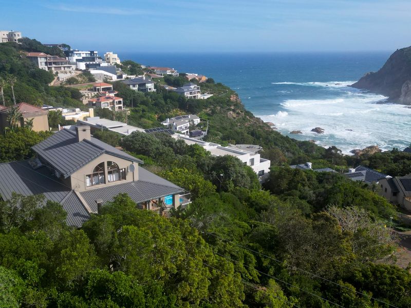 An elevated aerial view of a coastal residential estate featuring homes with modern pitched roofs, nestled in a lush hillside overlooking the ocean.