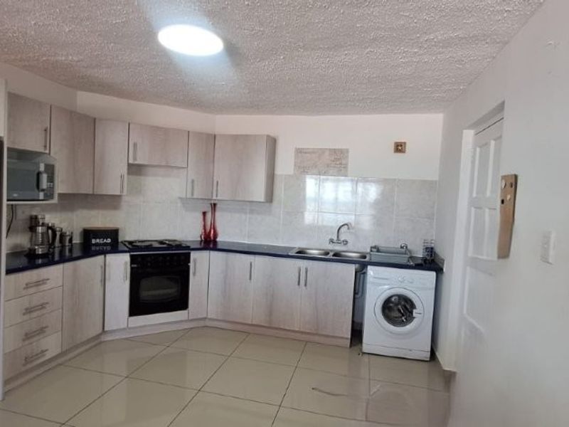 A functional kitchen featuring light wood-finish cabinetry, dark countertops, tiled flooring, and integrated appliances including an oven, hob, and laundry connection.