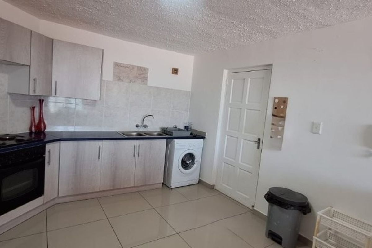 A functional kitchen featuring light wood-grain cabinetry, dark countertops, integrated oven and hob, tiled backsplash, and tiled flooring with an adjacent laundry connection.