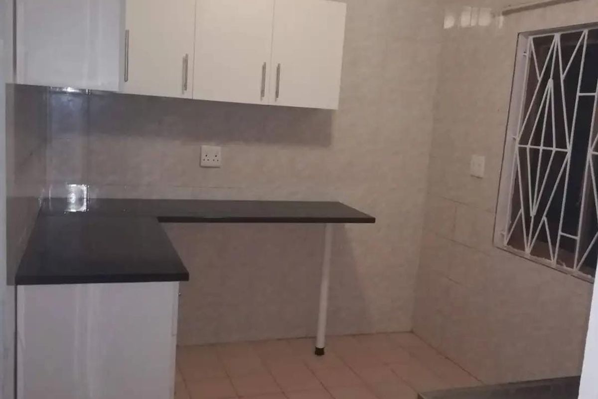 A functional, empty kitchen featuring white built-in cabinetry, dark countertops, tiled walls, and tiled flooring with a security-barred window.
