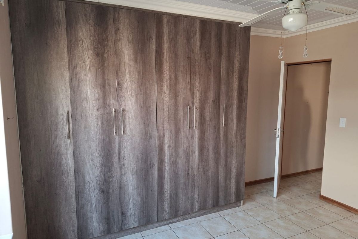 A spacious bedroom featuring extensive built-in cupboards with a modern wood-grain finish, tiled flooring, and a ceiling fan.