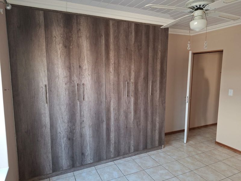 A spacious bedroom featuring extensive built-in cupboards with a modern wood-grain finish, tiled flooring, and a ceiling fan.