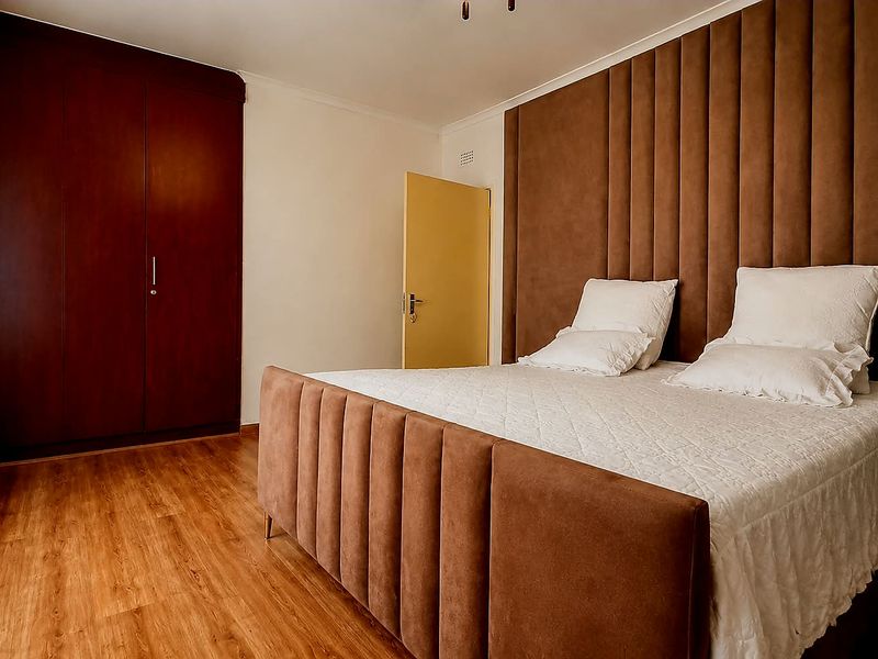 A well-maintained bedroom featuring laminate flooring, a large built-in wardrobe, and a decorative upholstered headboard wall panel.