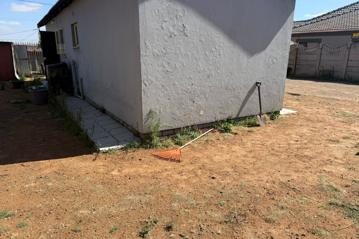 Rear exterior view of the property showing a plastered wall, tiled walkway, and surrounding unlandscaped yard area.