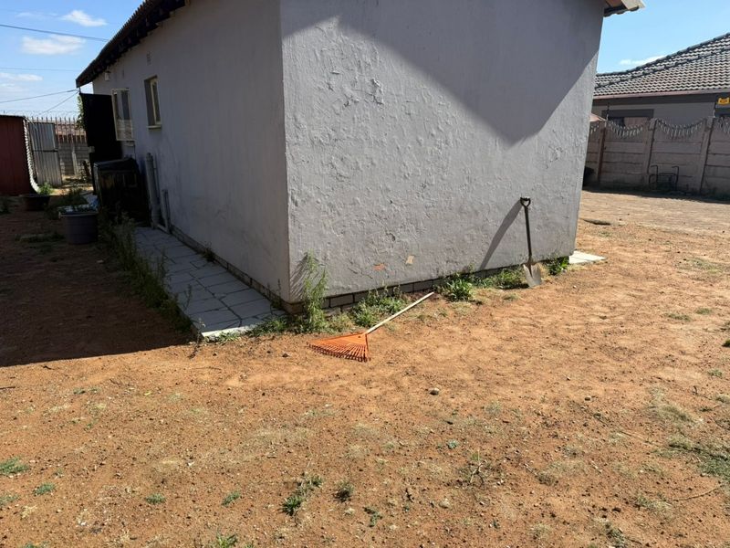 Rear exterior view of the property showing a plastered wall, tiled walkway, and surrounding unlandscaped yard area.