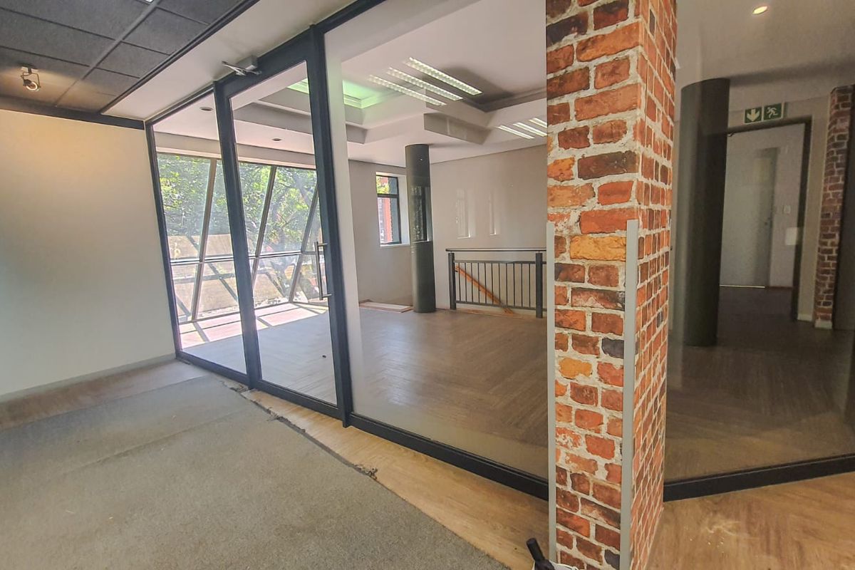 Open plan commercial office space featuring exposed brick columns, glass partitioning, and modern laminate flooring with a neutral aesthetic.
