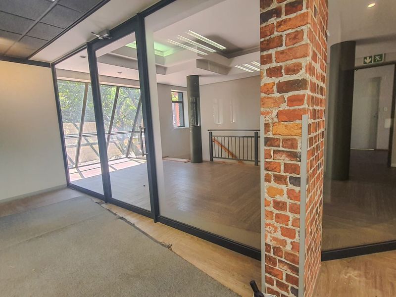 Open plan commercial office space featuring exposed brick columns, glass partitioning, and modern laminate flooring with a neutral aesthetic.